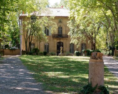 Mas du Jas de Bouffon of Cezanne in Aix-en-Provence. Warm stone 3-storey house with pitched red tile roof shaded by trees with green lawn in front and dappled view