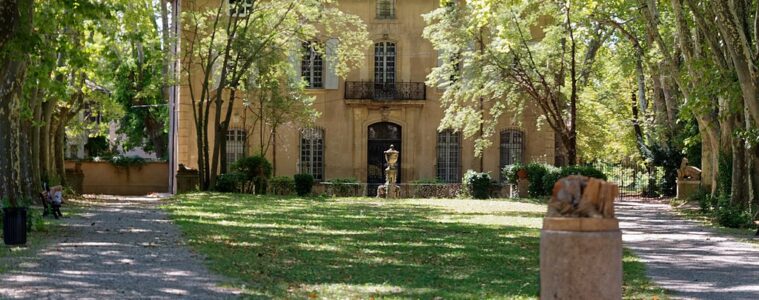 Mas du Jas de Bouffon of Cezanne in Aix-en-Provence. Warm stone 3-storey house with pitched red tile roof shaded by trees with green lawn in front and dappled view