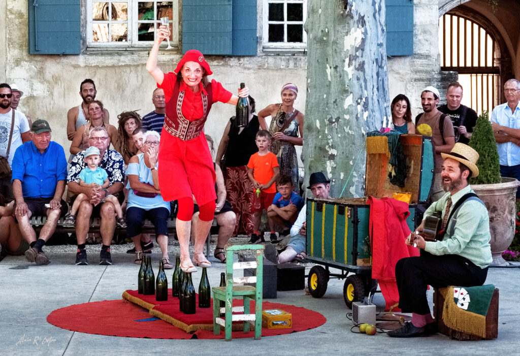 Font'Arts Festival with woman in red walking along series of wine bottles on ground holding a glass with small chair in front of her, man painting a pic of her and mystified onlookers