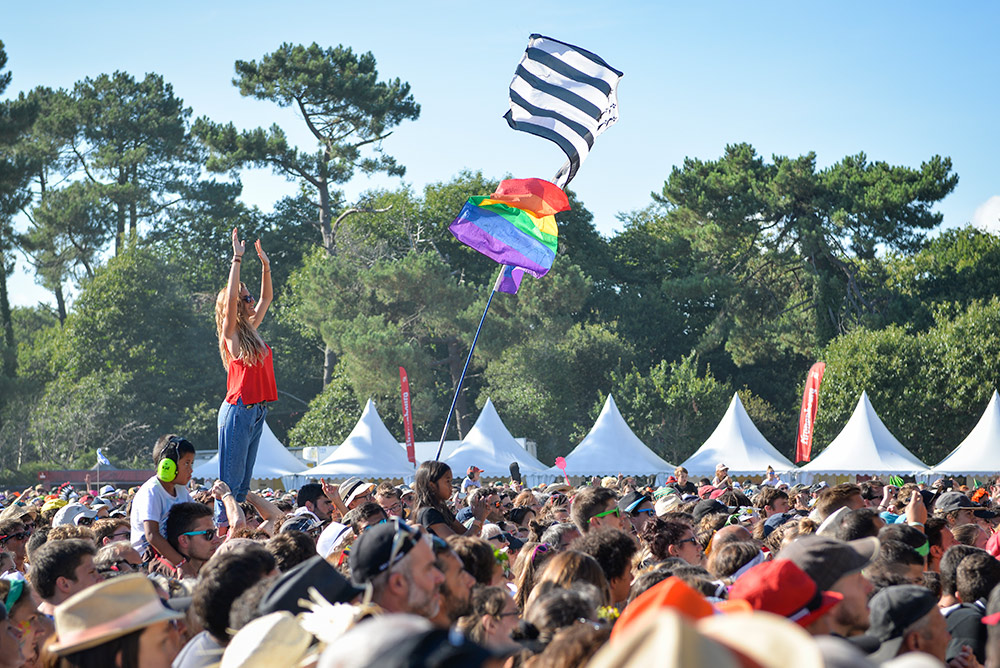 End of the World festival showing crowd in front with one woman in red top and jeans standing on shoulders so high above crowd, tents in background with gren trees and flags