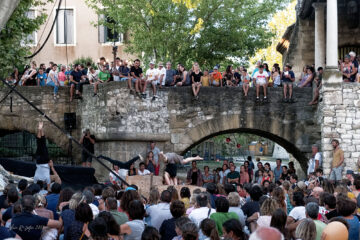 Font'Arts Festival with crowds on river bank and stone bridge watching 2 performers doing acrobatics on wooden blocks