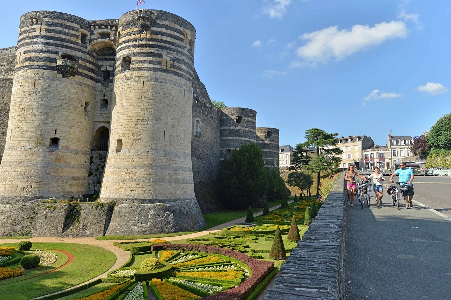 Angers castle looking fromone corner with street on right with people on bicycles and huge round tower and formidable walls on left with pretty gardens in front
