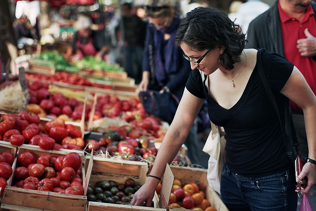 Woman in black top and jeans leaving over a stall picking out plus with boxes of peaches and apricots around
