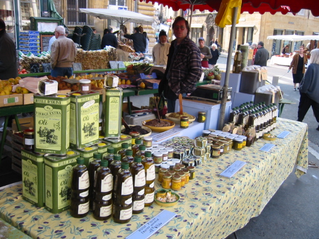 Stall at Avignon market with stall holdier behind and table with Probencal table cloth filled with bottles and cans of oliver oil