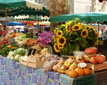 Very colourful stalls at Avignong market with green and white umbrellas above tables piled high with orange squash, sunflowers, garlic and more