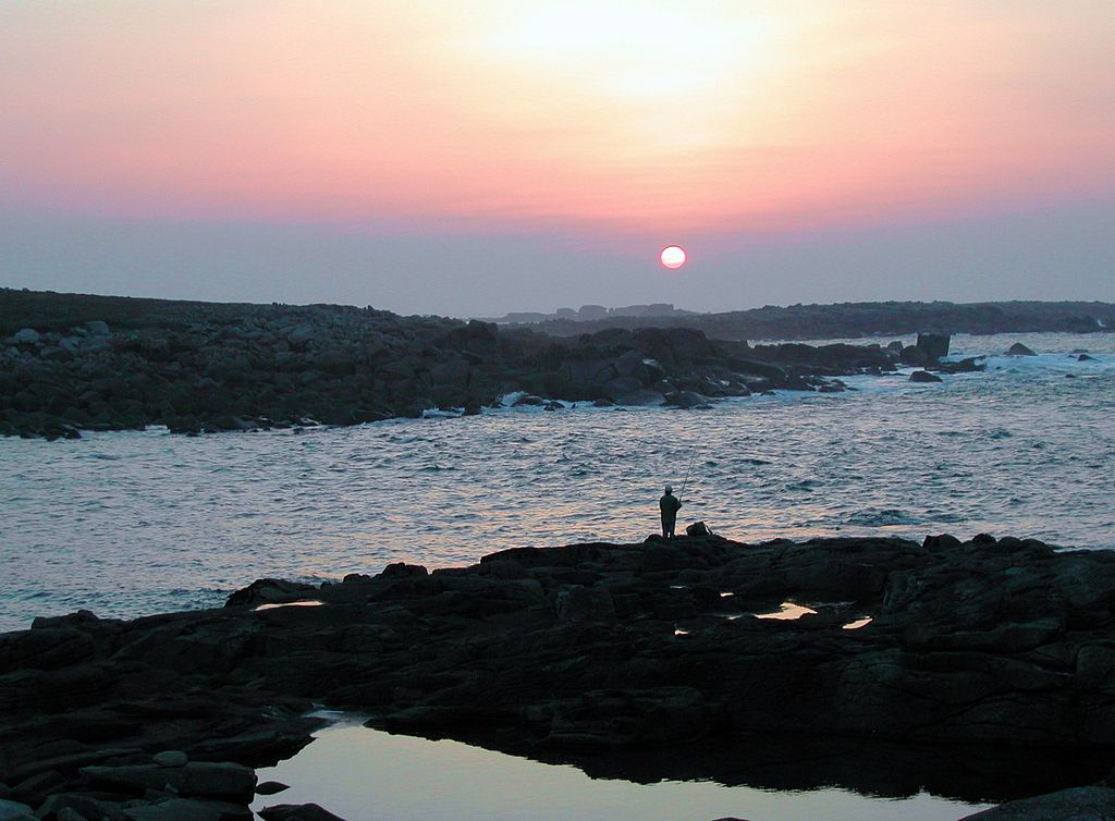 Sunset on Ile Grande Pointe de Toual with red sky, blue sea and rocks with lone small fisherman