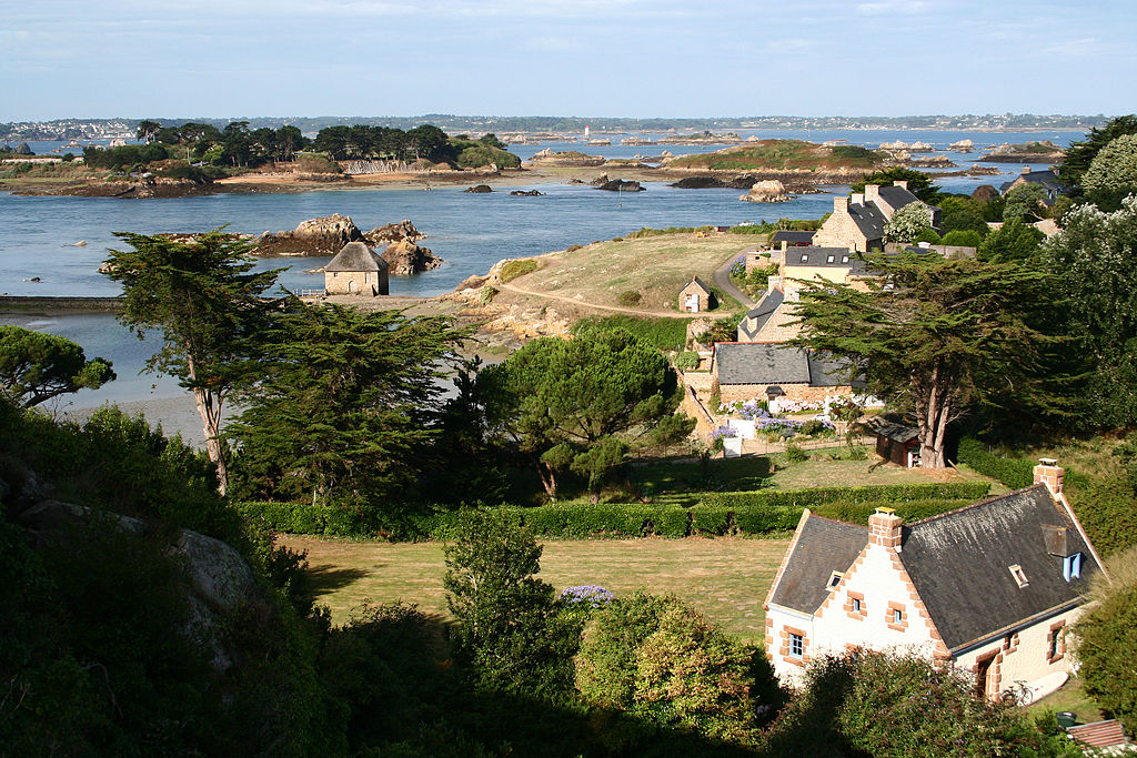 View from high up over small houses of stone, fields and trees and sea in background in bay