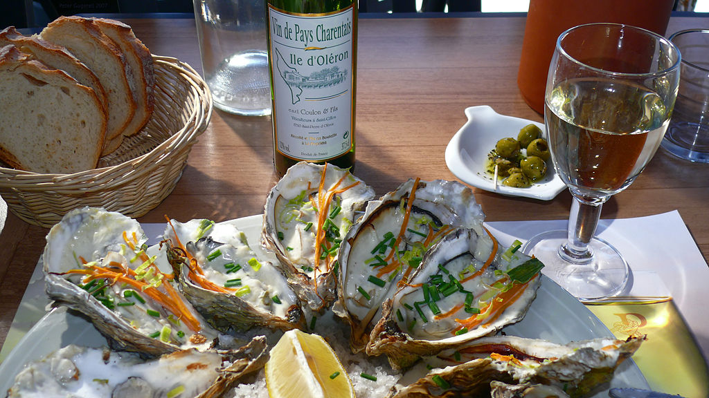 Food shot of oyster in shells with green parsleyand lemon in front with bottle of Oleron wine behind and other small dishes