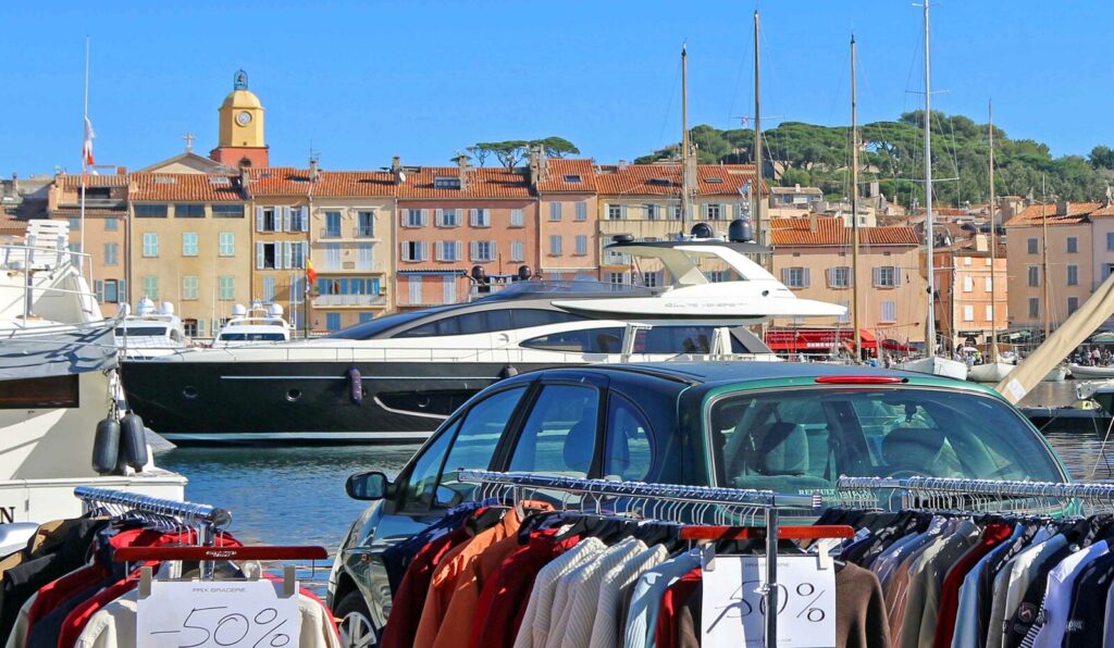 Grande Braderie of Saint Tropez with shops putting rails of clothes outside. This one from shop showing clothes railes, a car in front of the water and yachts and houses behind