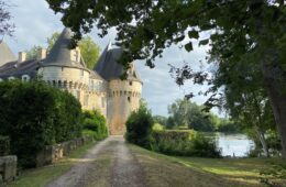 Chateau de Bazouges Loir Valley. Path beside green lawn and river to right to turreted castle in distance in sunlight