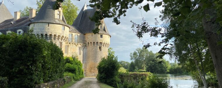 Chateau de Bazouges Loir Valley. Path beside green lawn and river to right to turreted castle in distance in sunlight
