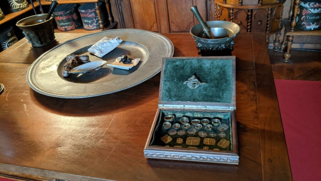 Bauge apothecary showing close up of wooden table with pewter dish with papers scattred on it, pewter pestle and mortar behind and shallow iron box with shapes that look like sweets
