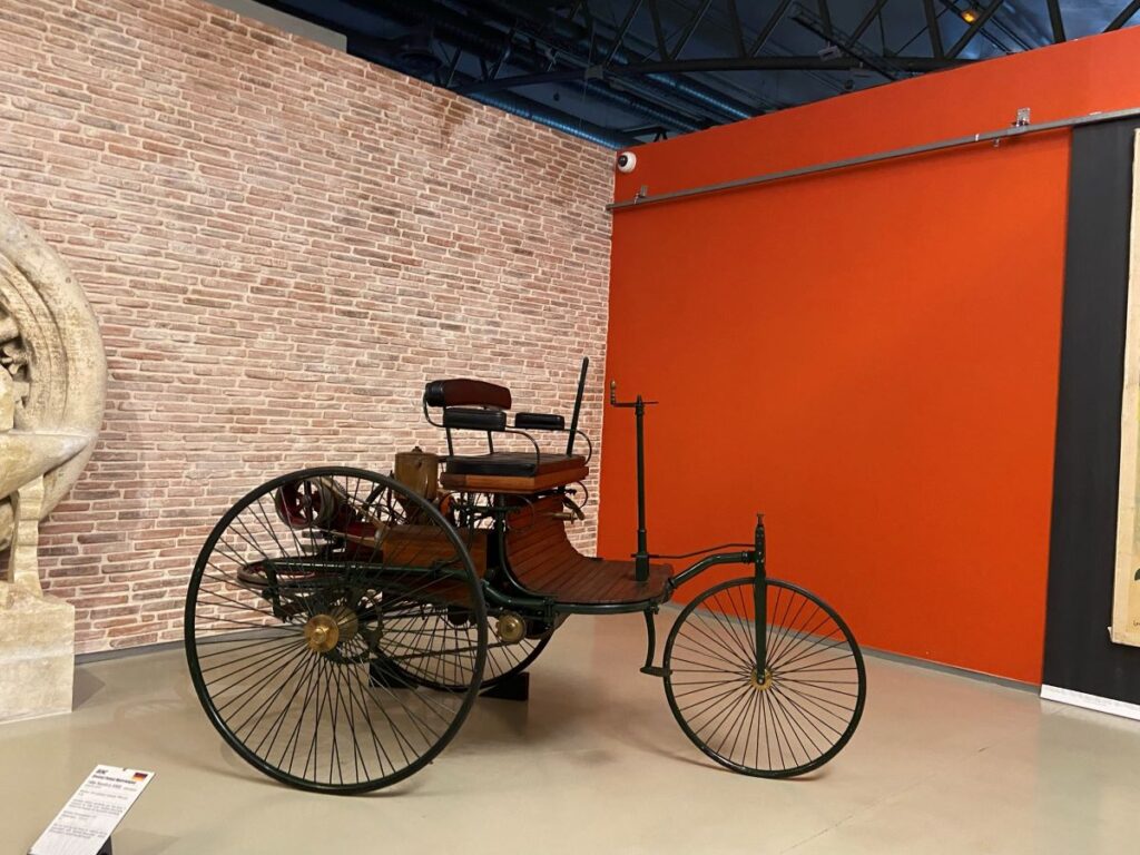 Replica of first Benz car a 3-wheeler with large wheels behind and small in front and seat and steering gear looking more like a bicycle