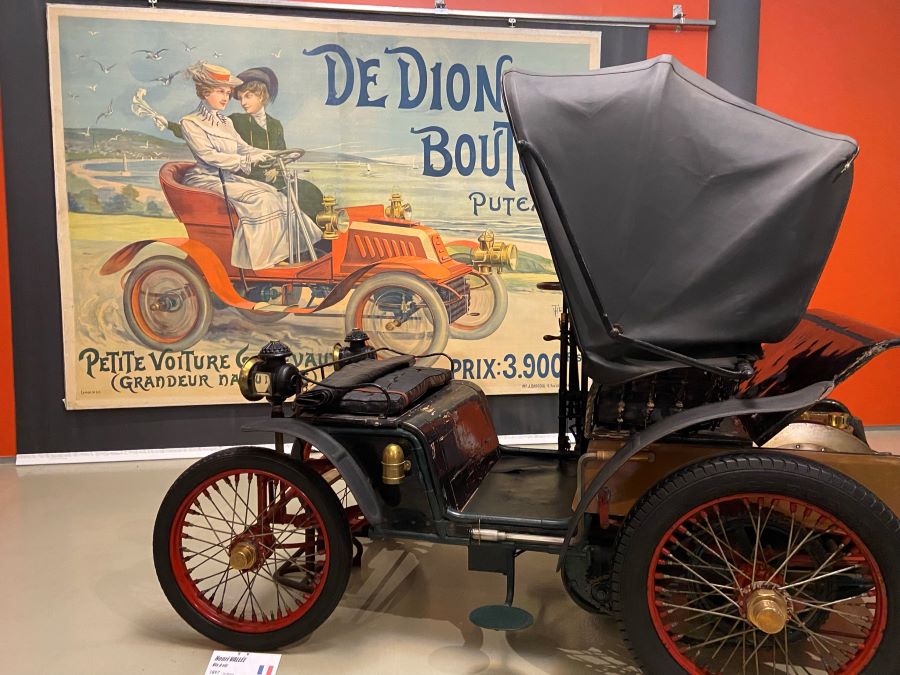 De Dion Boulton car with hood up in front of poster showing early 20th century ladies in their fab car