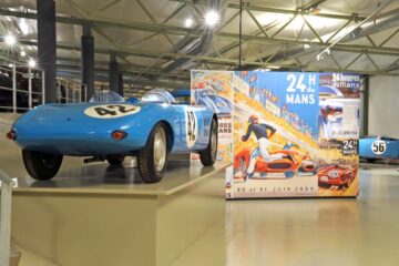 Museum of the 24 hours of Le Mans showing old blue racing car and big poster behind advertisin gthe race with old photo in hangar like building