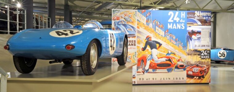 Museum of the 24 hours of Le Mans showing old blue racing car and big poster behind advertisin gthe race with old photo in hangar like building
