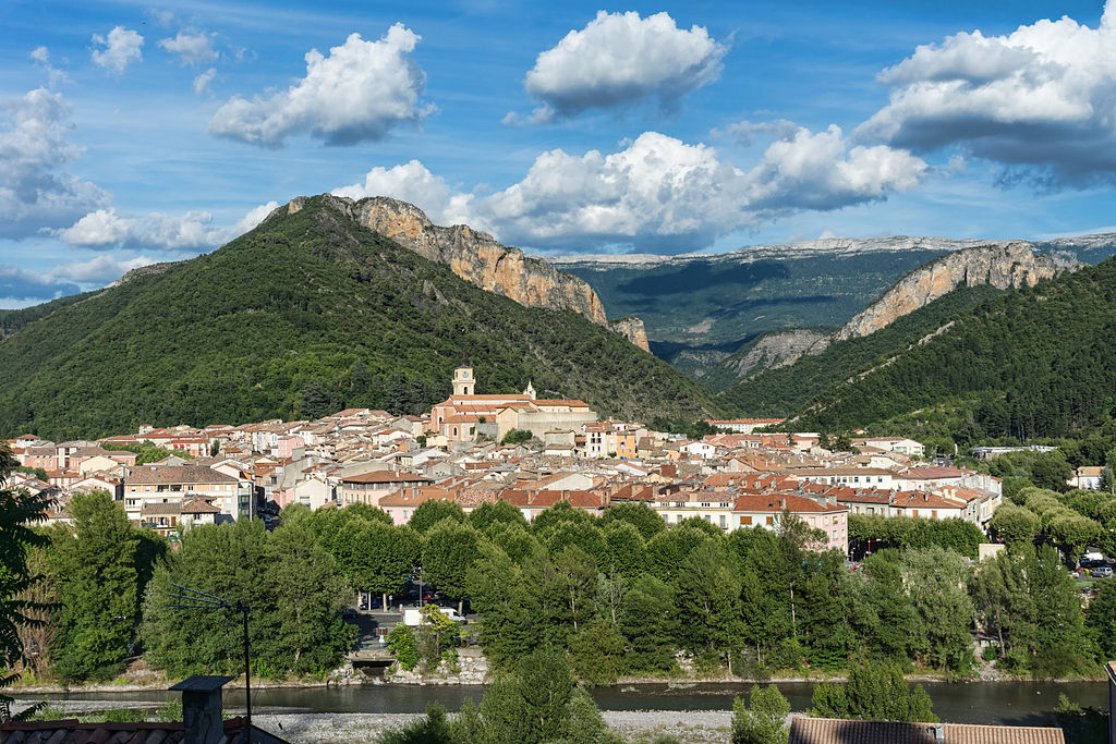 View from afar of Digne-les-Bains with mountains i the background, some wooded, some bare and village spread out on hillside below wich church towers at top of the town