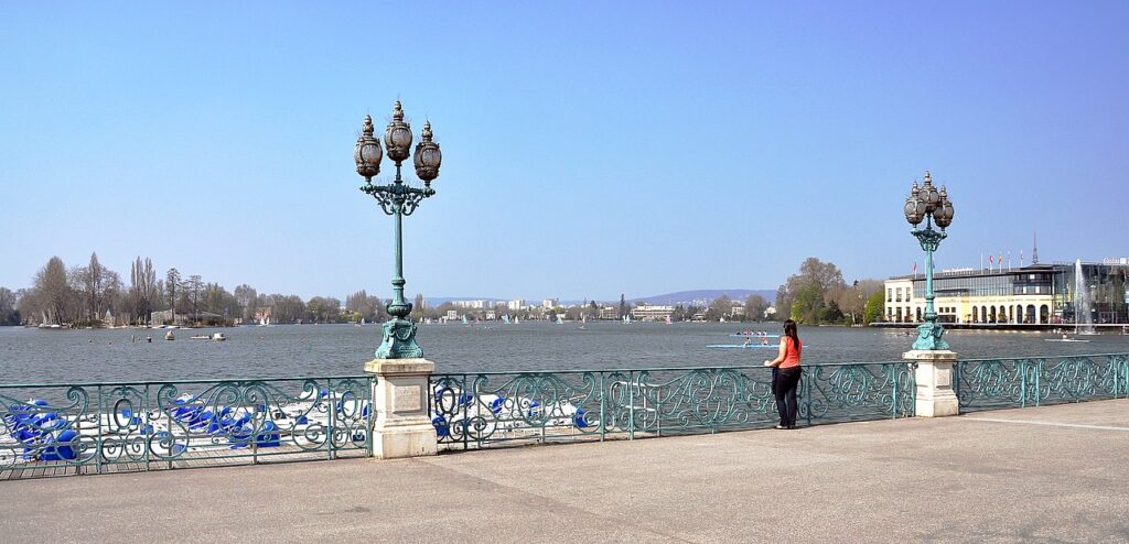 Lake at Enghien les Bains, suburb of Paris. Woman standing near ornate pillar with three lights on top looking out at blue lake with buildings in distance