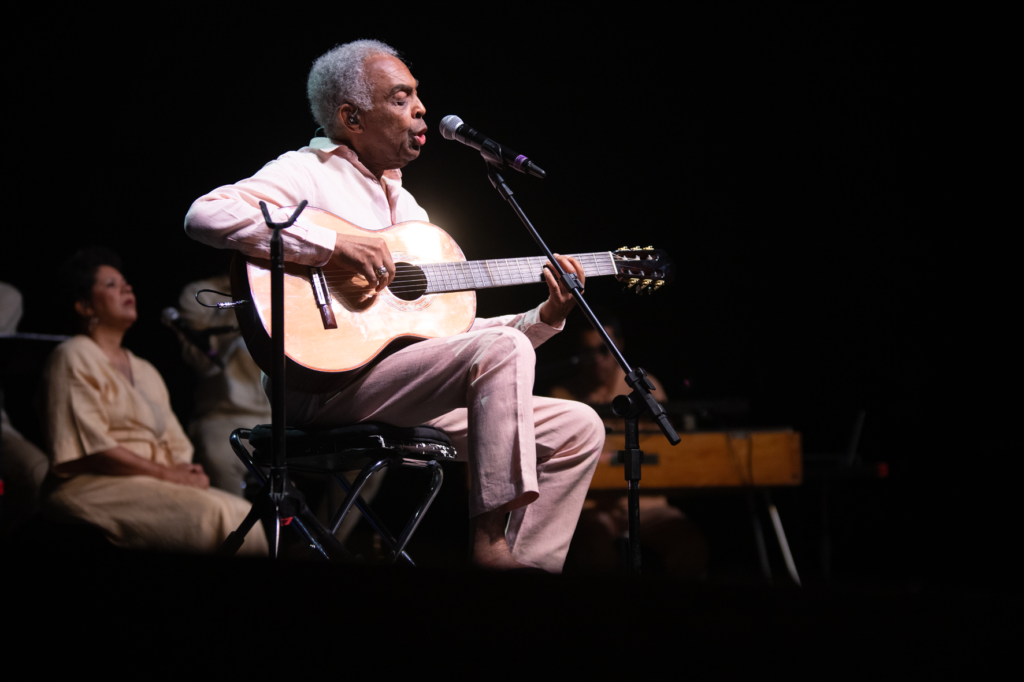 Gilberto Gil on stage at Marciac Jaz Festival playing a guitar, seated, and singing into microphone