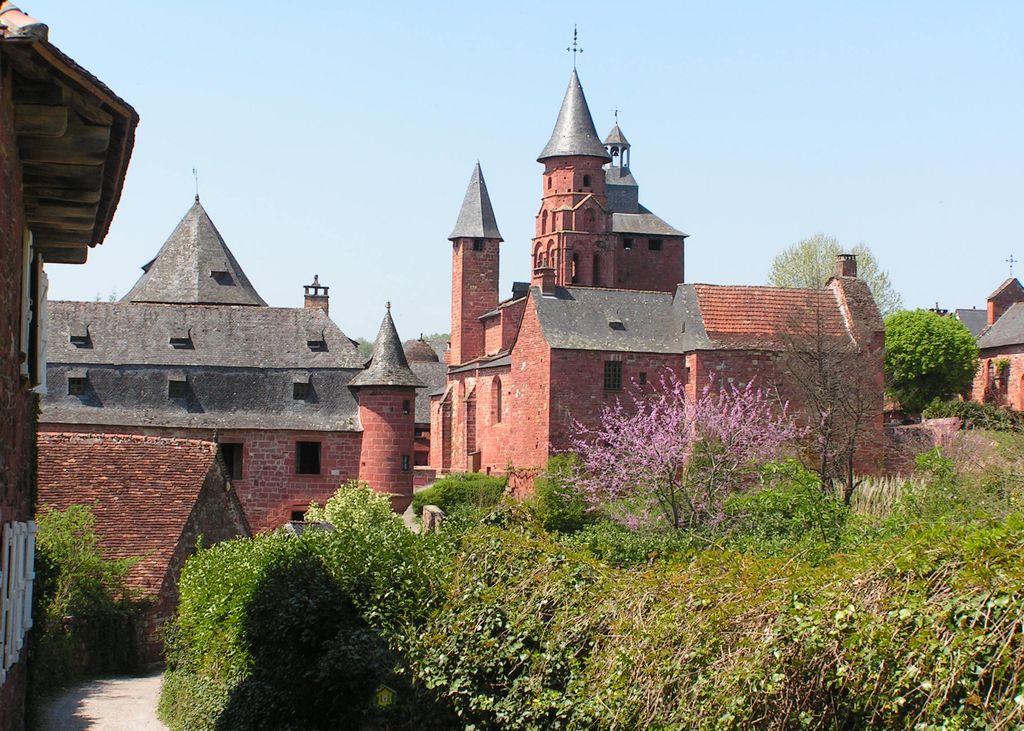 Collonges-la-Rouge village showing bushes at front with red stone houses with towers and conical tops behind