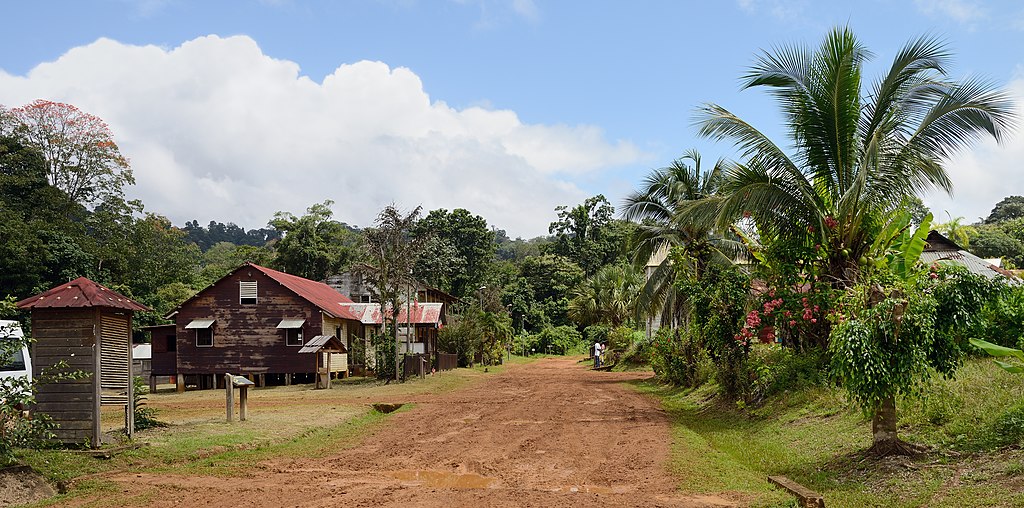 Dirt track in Saul in French Guyana with red soil and houses on left with corrugated iron rooves and trees