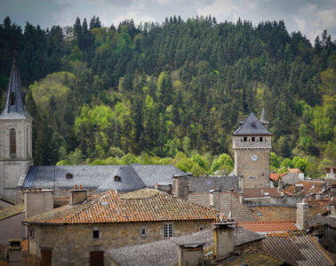 Looking over red tiled rooftops with round and square towers of Le Malzieu with wooded hills behind