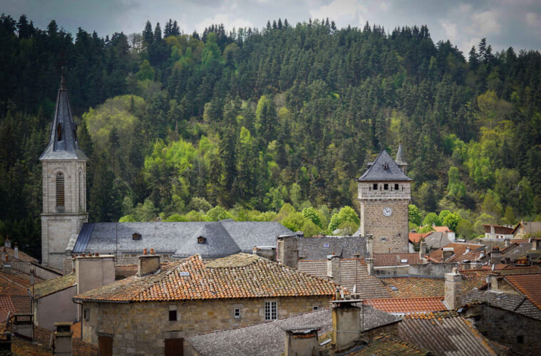 Looking over red tiled rooftops with round and square towers of Le Malzieu with wooded hills behind