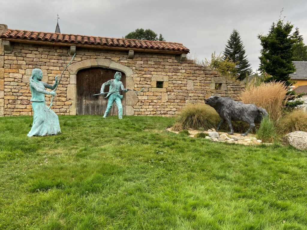 Old barn withclosed doors in front of green patch with two bronze figures with pitchforks holding the bete du gevaudon at bay