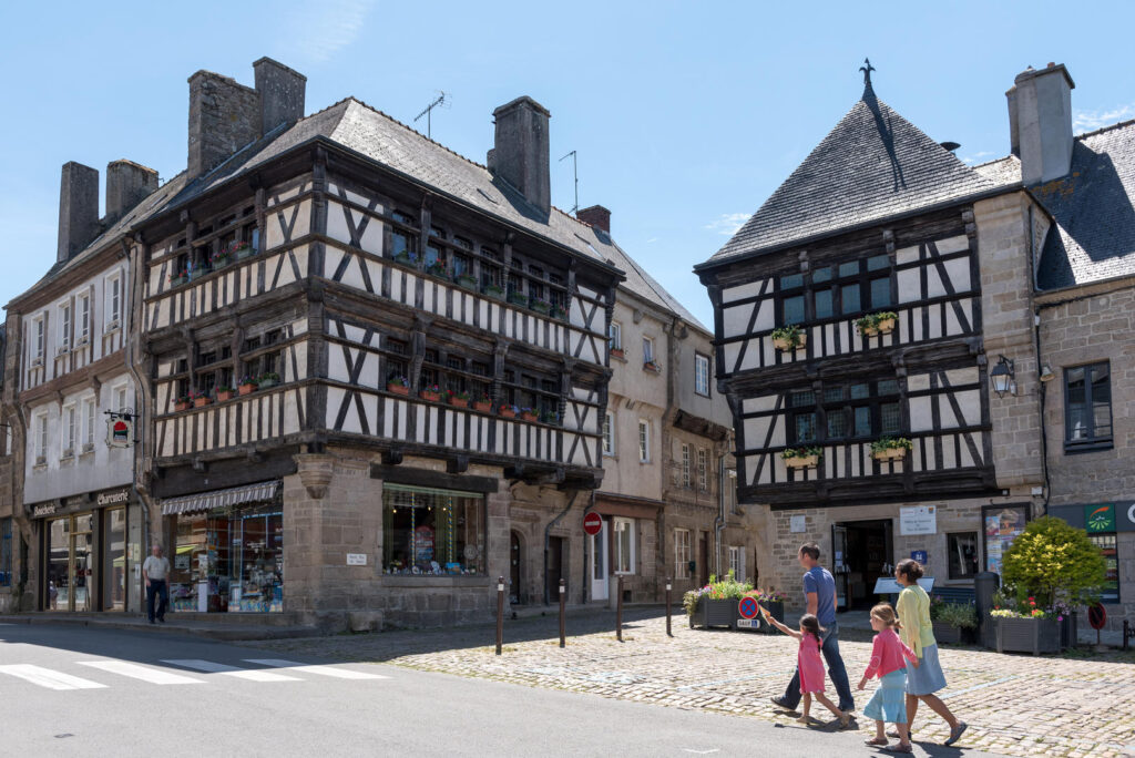 Quinton village in Brittany with old stone streets and familyon left walking past high 3-storey houses with sloping rooves and half timered elaborage pattern on 2 upper floors and stone with shop windows on ground