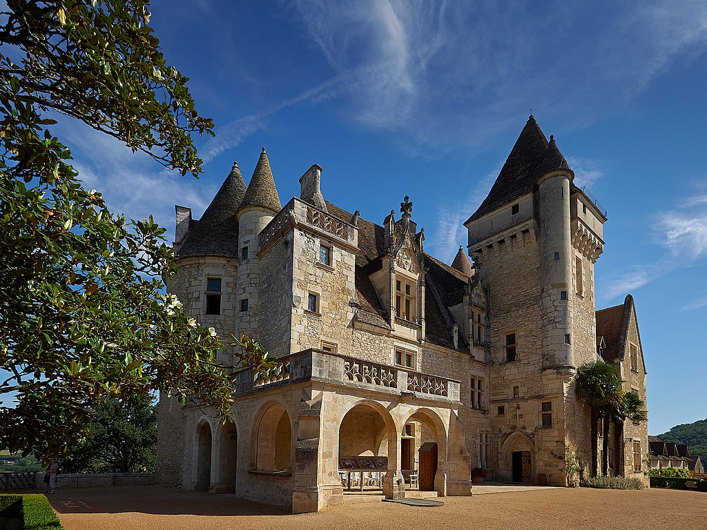 Chateau des milandes from front showing large tower to right, rooves, walls and stone balcony in front of gravel drive with tree to left