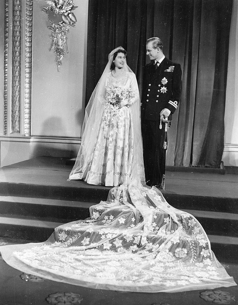Black and white photo official portrait of Princess Elizabeth and Philip Mountbatten at their wedding on steops looking at each other with long train of her dress cascading down steps