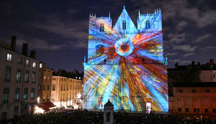 Lyon cathedral facade litup for festival with lights mainly shandes ofblue and red radiating from central rose window at night with crowds in front