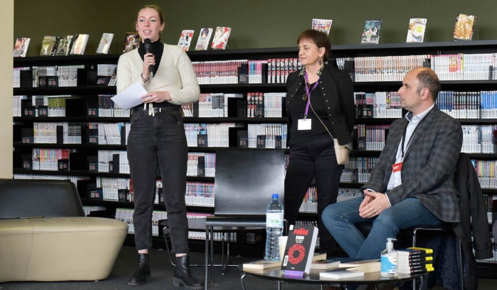 Sylvain Forge at Lyon Crime festival showing her on stage with microphone and woman beside her and shelves of books behind