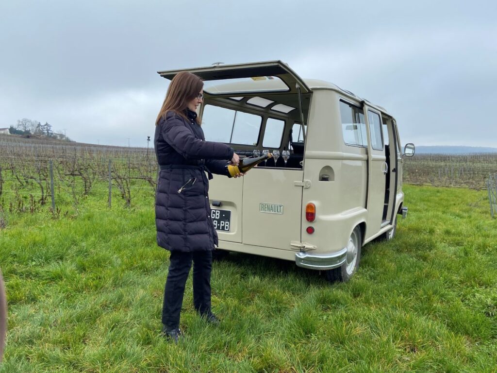 Woman standing in front of back of vintage van holding bottle of champagne and sabre as she beheads the champagne bottle