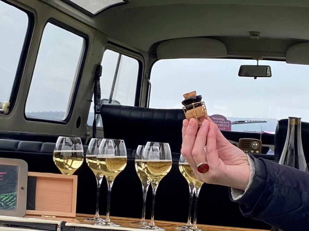 Hand holding beheaded champagne cork and bottle neck in front of van with back down and glasses of champagne