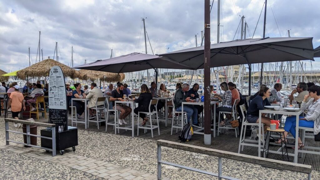Les Sables d'Olonne Les Patagos restaurant view from one sideof street showing parasols and people on chairs at tables eating with harbour with yachts behine
