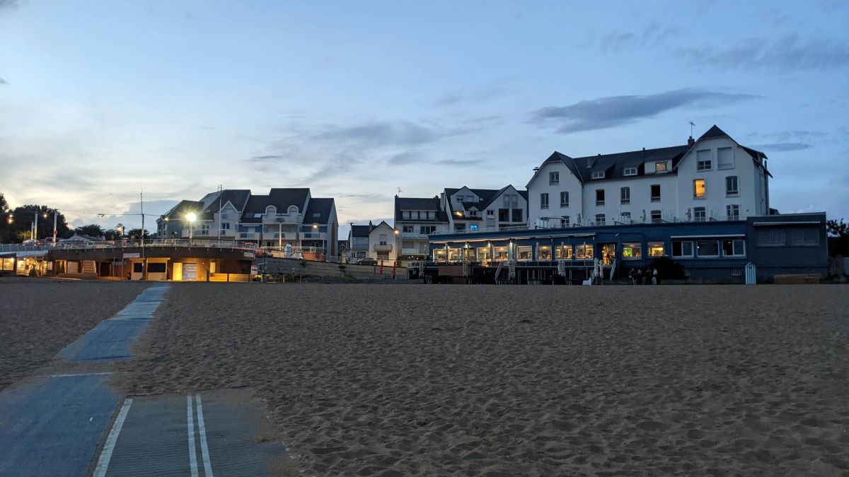 Monsieur Hulot's hotel seen from beach. Long sandy beach in front and one large white hotel building with terrace and tables outside with another large white building beside