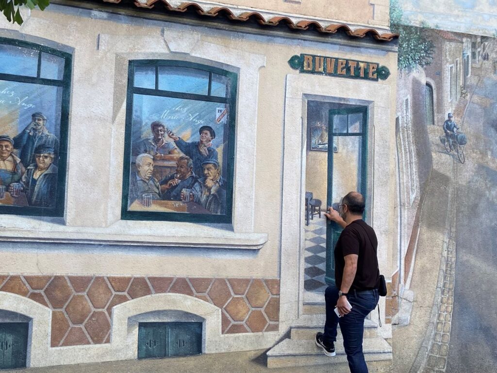Mural of a buvette (bar) on wall with real man trying to ope the door of the buvette and figures seen in window drinking