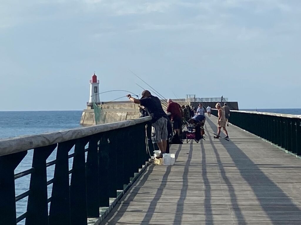 Long boardwalk on stone jetty with fishermen on one side and at end red lighthouse