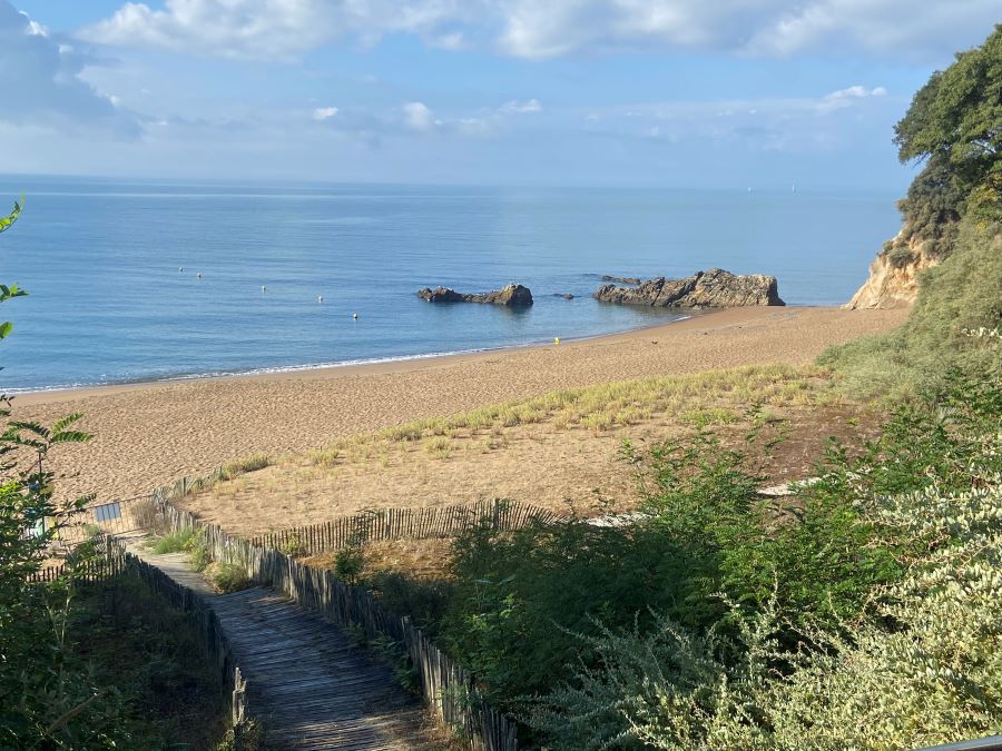 Smugglers Path in Saint-Nazaire looking down from top path with shrub to steps down to long sandly beach with sea in background and blue sky