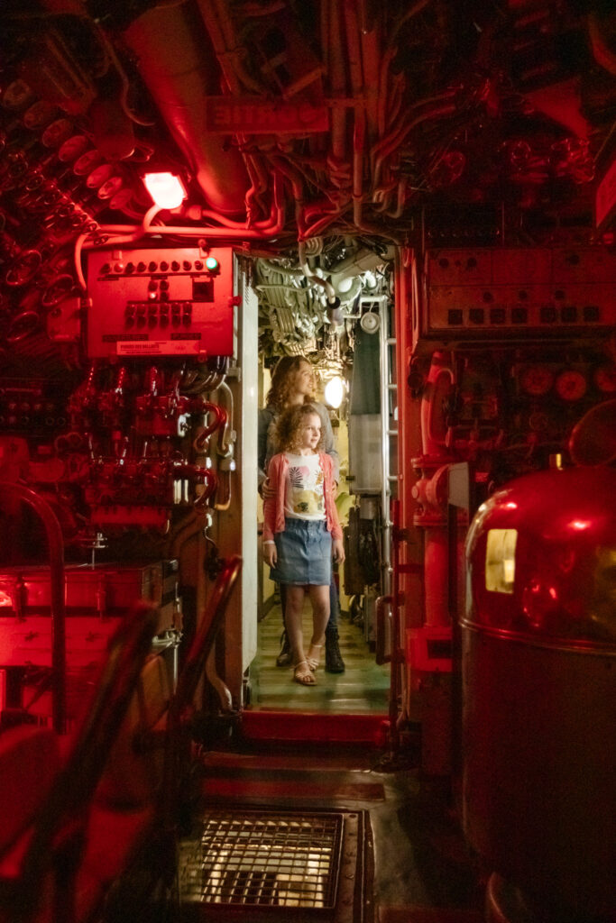 inside espadon submarine museum in saint nazaire showing narrow corridor in orange light with girl and parent walking towards camera. All sorts of equipment on either side