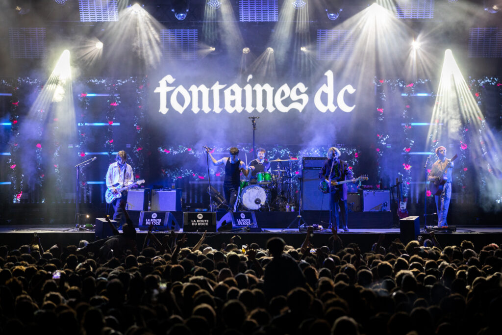Fontaines at la Route du rock showing rock band on stage lit by blue lights, audience in front and name fontaines de above