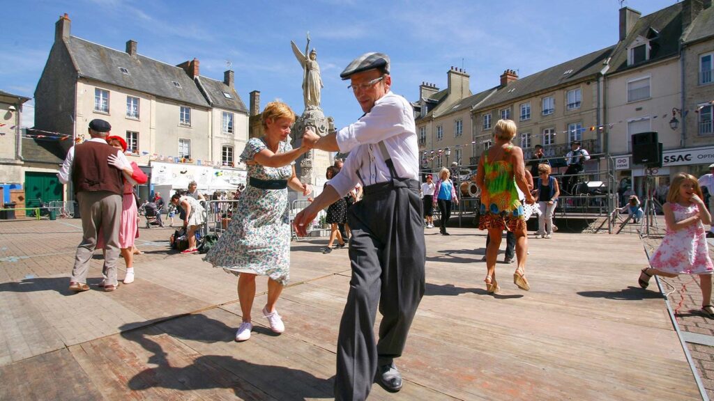 Couple in 1940s costume dancing in square at Carentan with statue behind and old houses