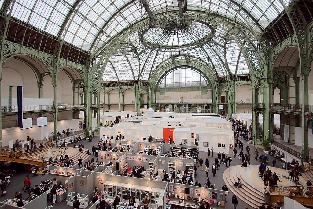 Paris photo show shot from top of Grand Palais showing huge iron and glass Victorian roof ovr large space full of booths with many people and stage in background