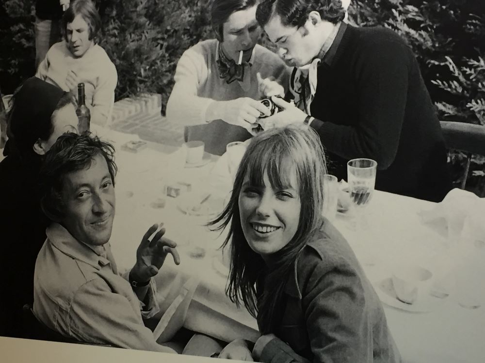 Jane Birkin and Serge Gainsbourg at table with other figures behind black and white