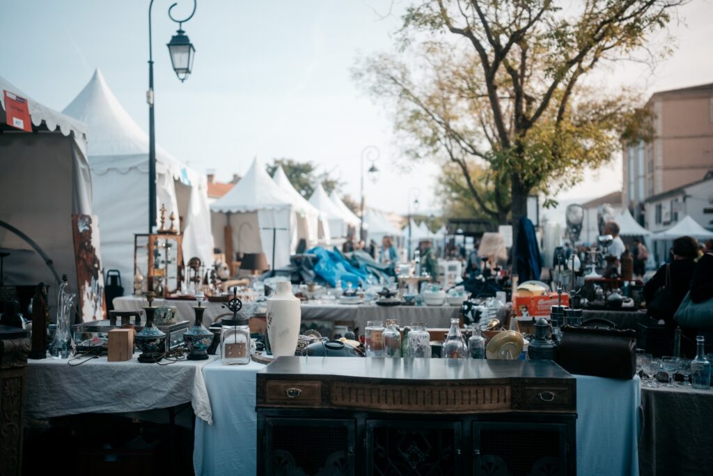 L'isle sur la Sorgues antiques market with photo taken from behind table piled high with small antiques looking out onto square with white pavilion tents with more stalls