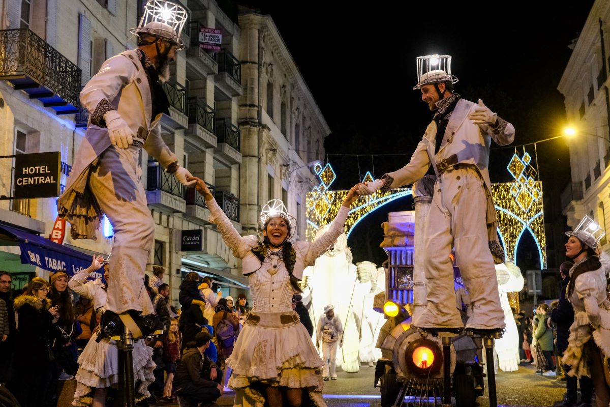 Night at Avignon Christmas Market with people in street, two very tall men holding girl between them in middle, all in old medieval costume