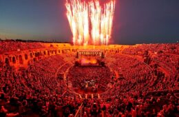 Nîmes Festival with huge crowd around circular stage bathed in organse glow from fireworks in background