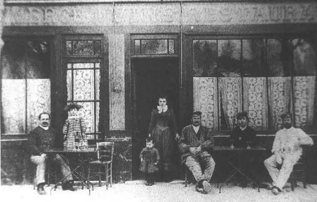 Black and white photo of Auberge Ravoux where van Gogh spent his last days. Exterior front of wine shop with one laady standing and men sitting at outside tables