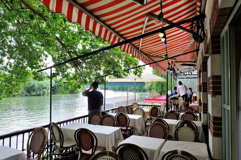 Chatou Fournaise restaurant beside the Seine with river on left, tables and chairs on a terrace and a red and white striped awning above
